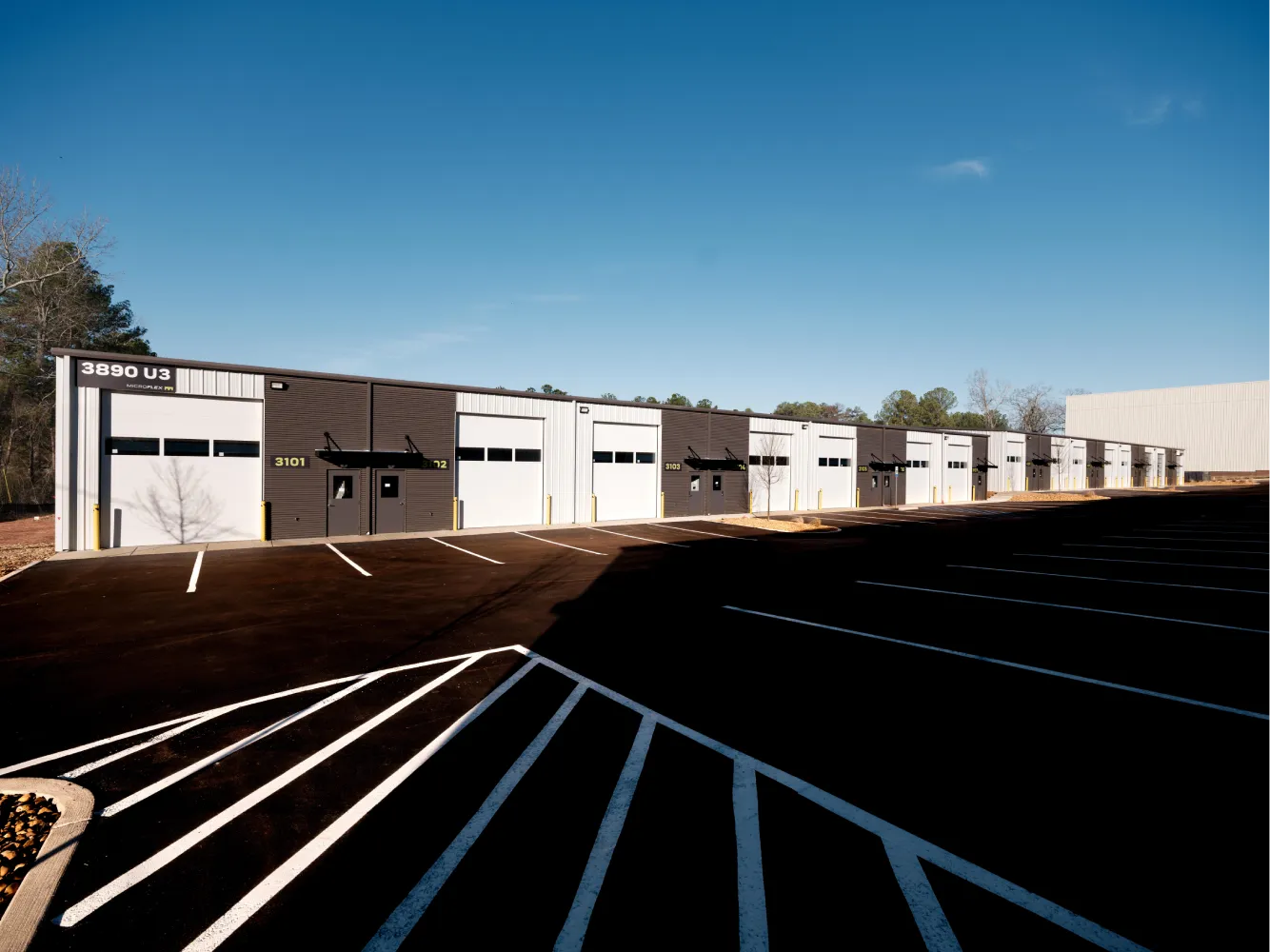Exterior row of storage unit bays with roll-up doors, brown and white panels, and a large parking lot under a clear blue sky.