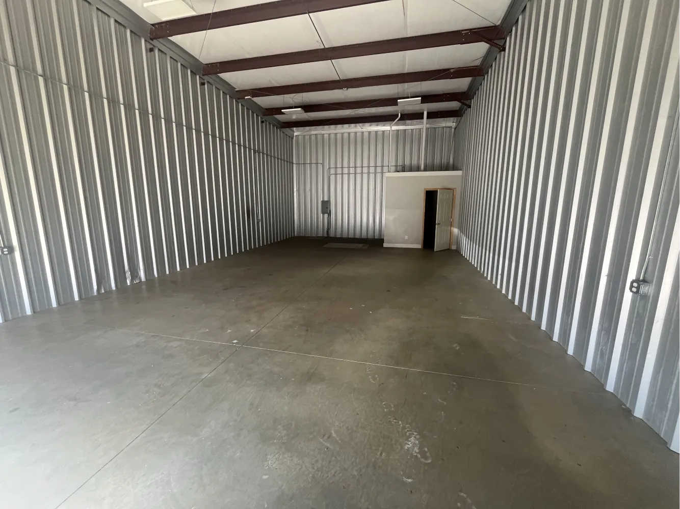 Empty corrugated metal-walled warehouse interior with exposed steel beams, concrete floor, and back room with a white door.