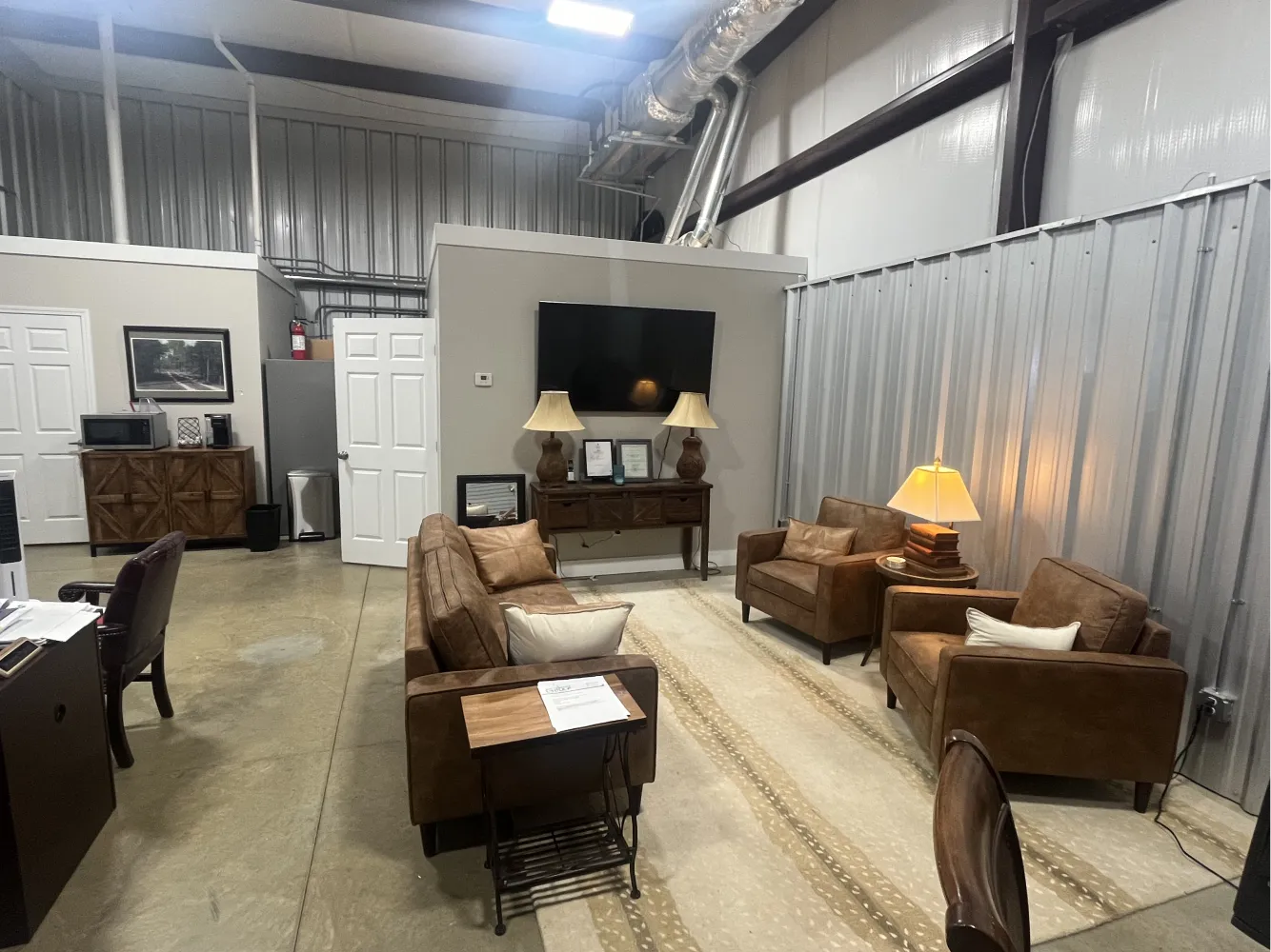 Industrial warehouse lounge with brown leather seating, beige rug, wall-mounted TV, lamps, and exposed metal walls.