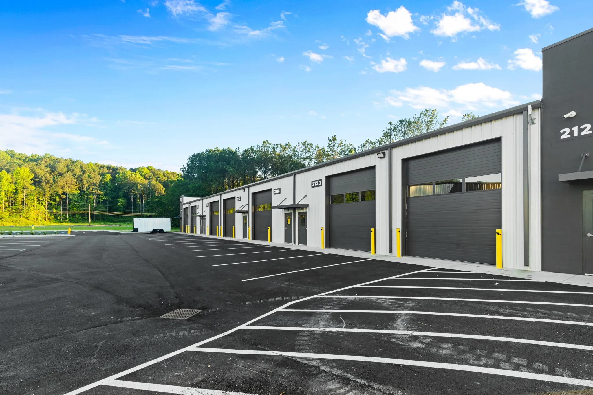 Row of gray warehouse units with roll-up doors along a paved parking lot, yellow bollards, and blue sky.
