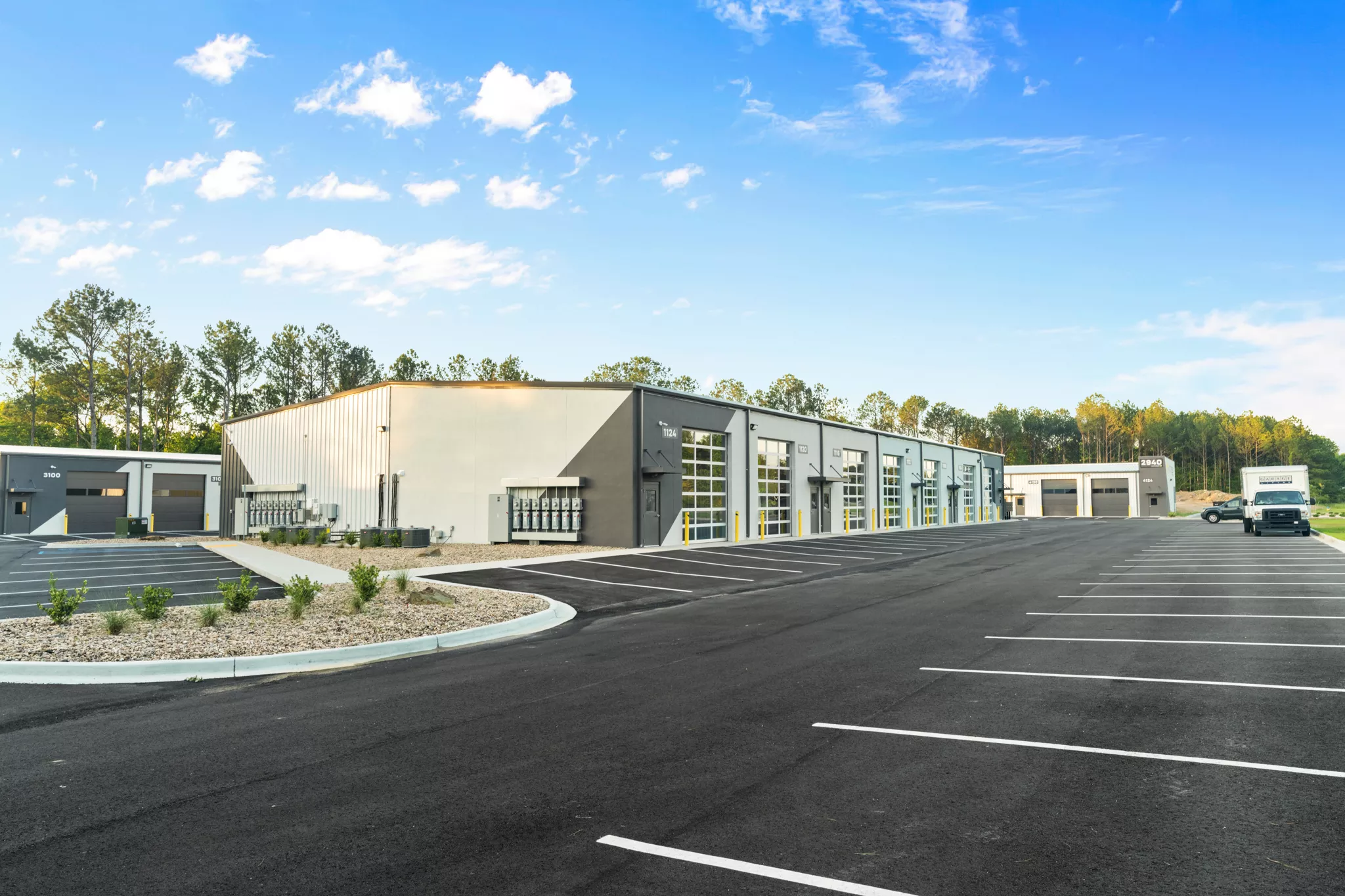Row of modern flex warehouse units with roll-up doors and glass entrances, plus a large parking lot under a blue sky.