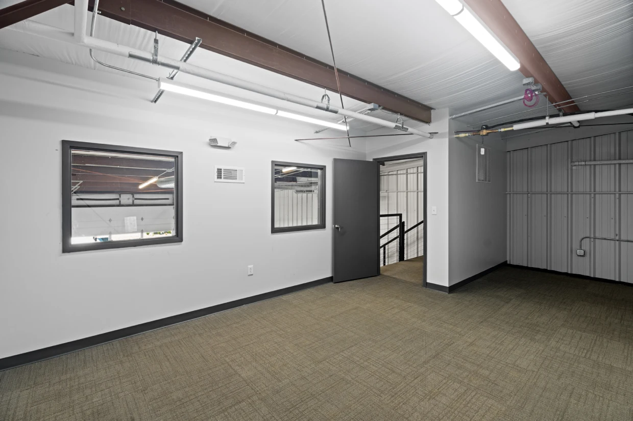 Interior of a small flex space with two windows, doorway to a stairwell, exposed ceiling pipes, carpeted floor, and a gray corrugated metal wall.