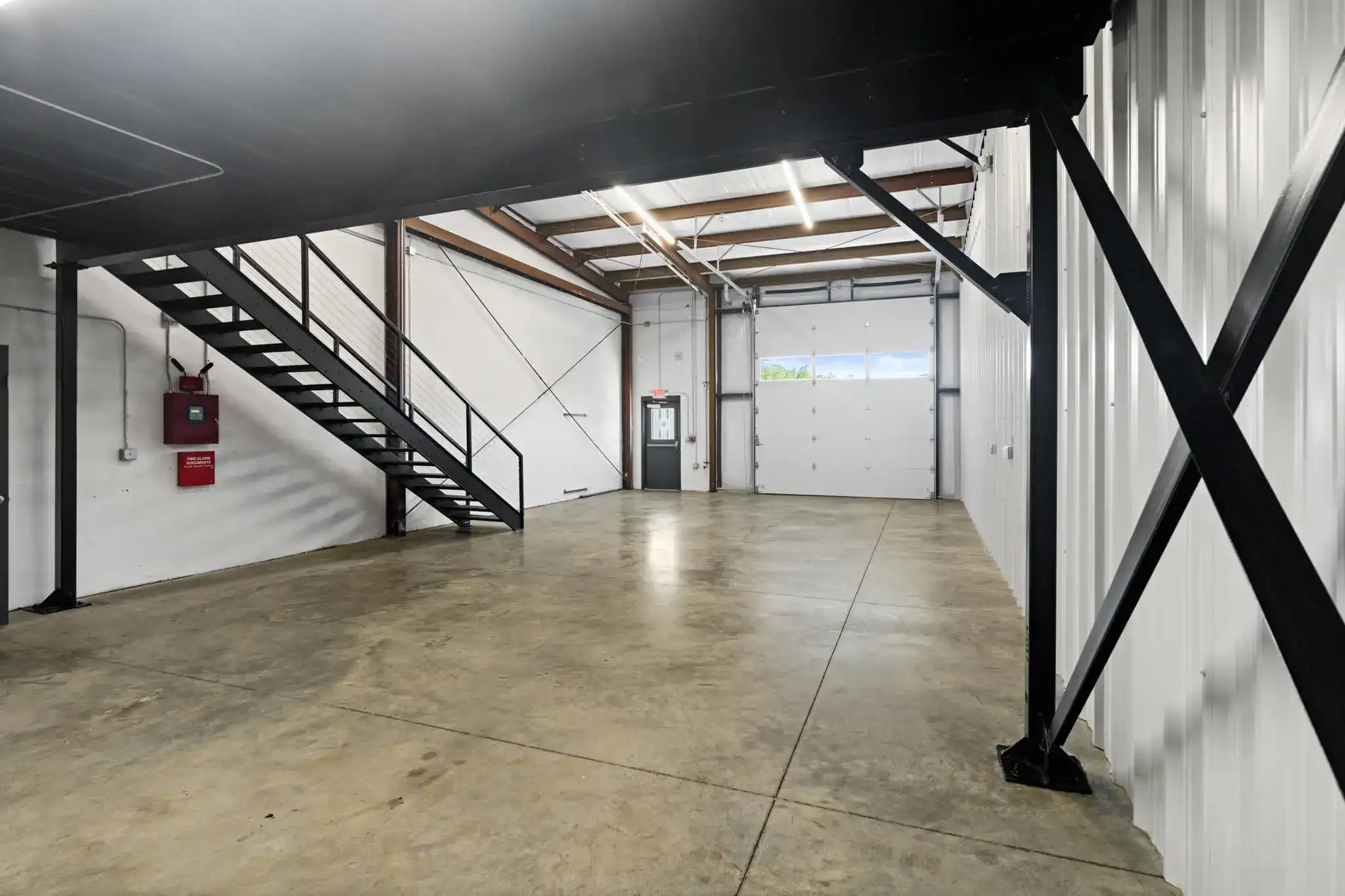 Industrial warehouse interior with concrete floor, black metal mezzanine stairs, white walls, and a roll-up door.