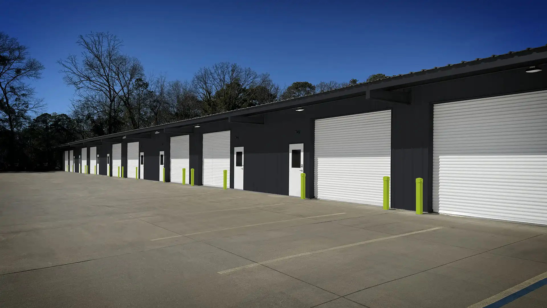 Exterior view of a row of storage units with white roll-up doors, gray walls, green bollards, and a concrete parking lot.