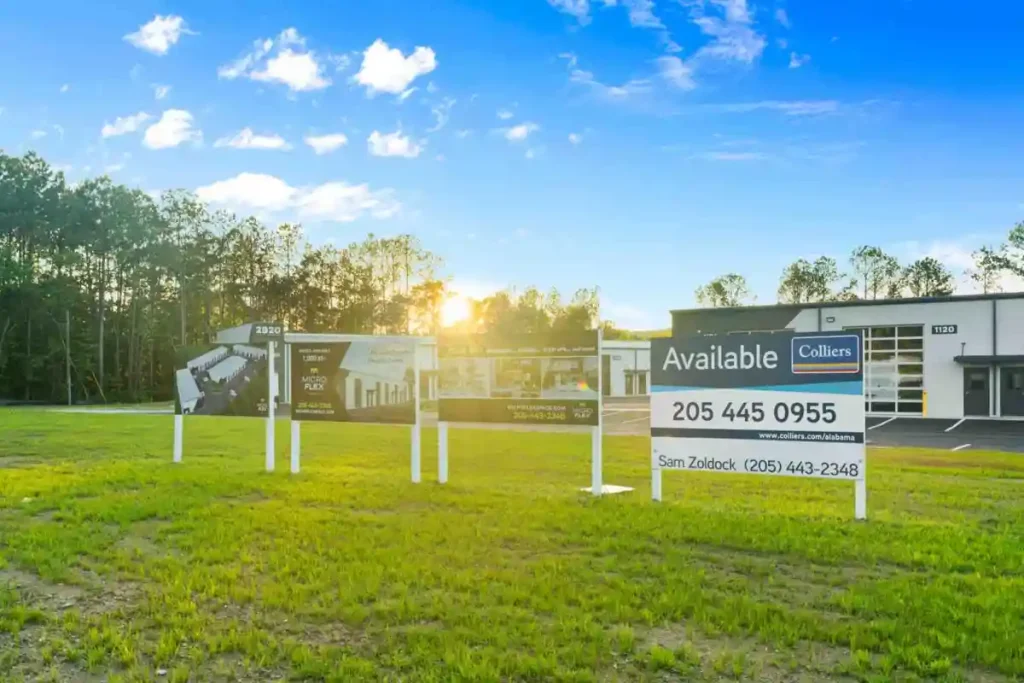 Row of available real estate signs on posts in a grassy lot with a modern commercial building in the background.