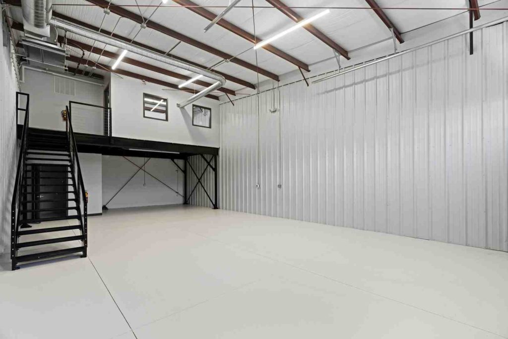 Spacious industrial warehouse interior with corrugated metal walls, a black metal staircase to a mezzanine, exposed ductwork, and bright overhead lights.