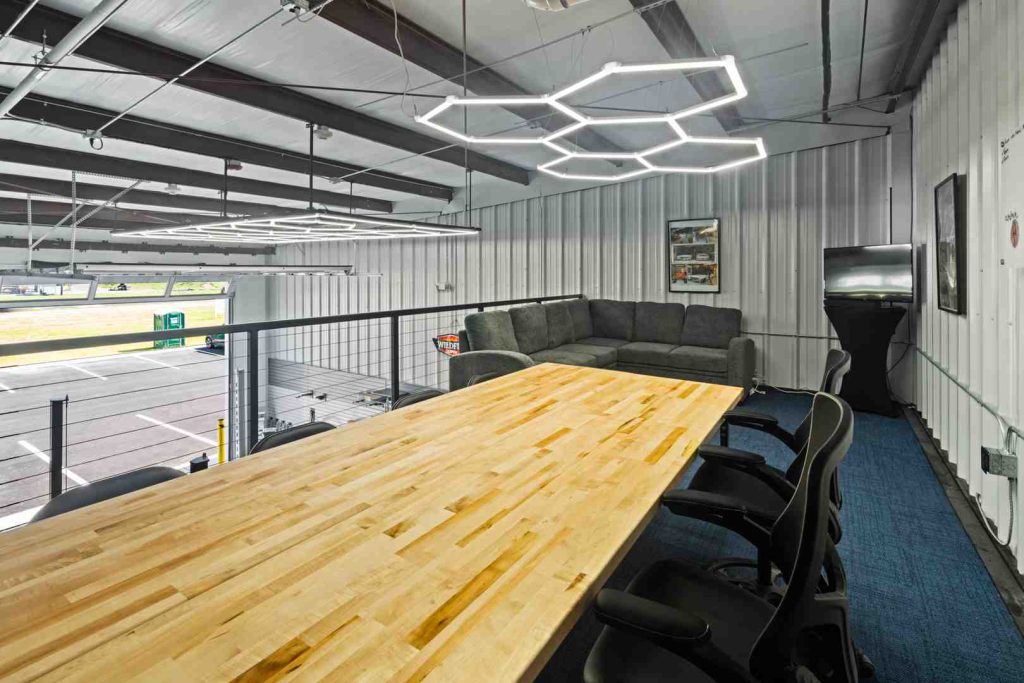 Mezzanine office space with a long wooden conference table, black chairs, a gray sectional sofa, hexagonal LED lights, and a view of the parking lot below.