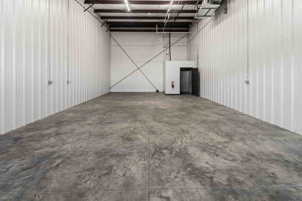 Wide view of an empty industrial warehouse interior with concrete floor, white corrugated walls, ceiling lights, and a back room with a door.