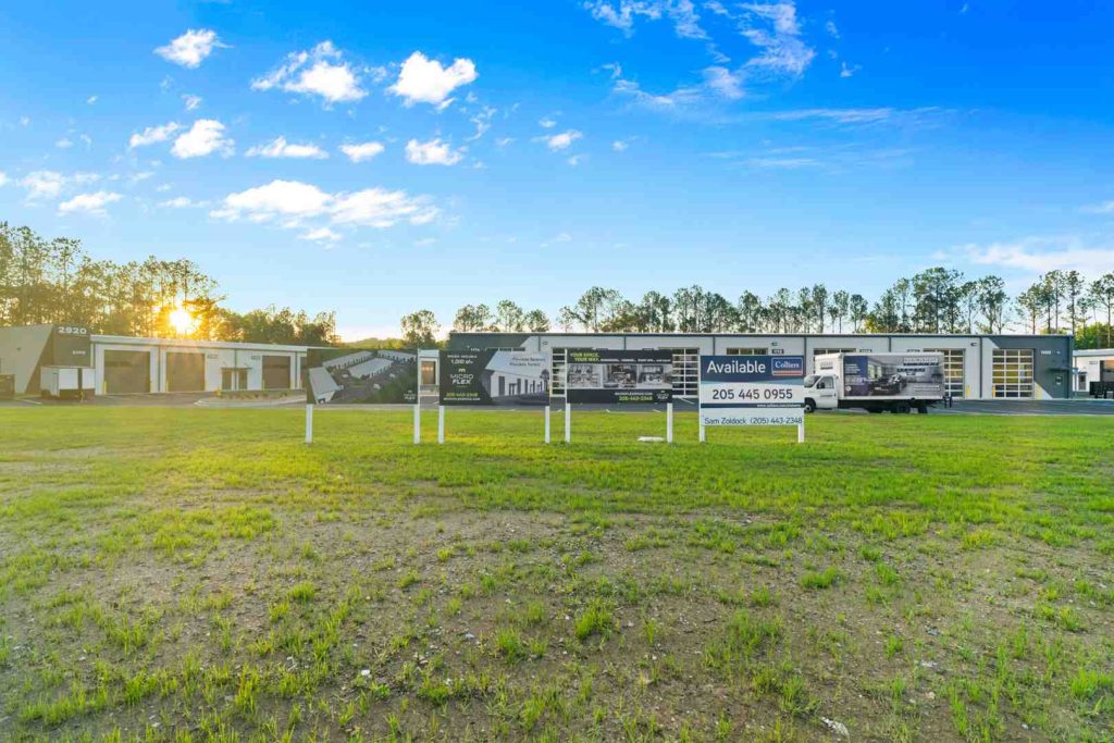 Sunlit row of modern flex warehouse units with roll-up doors and advertising signboards along a grassy lot.