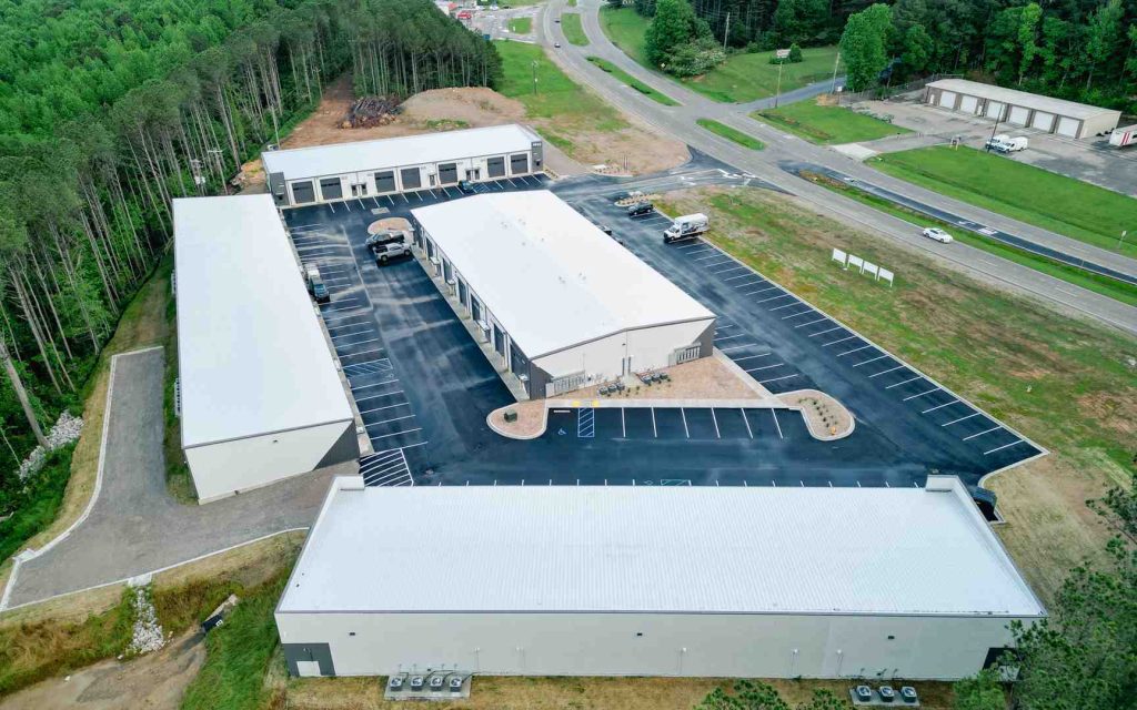 Aerial view of a modern warehouse complex with long central building, loading bays, extensive parking, and surrounding trees.