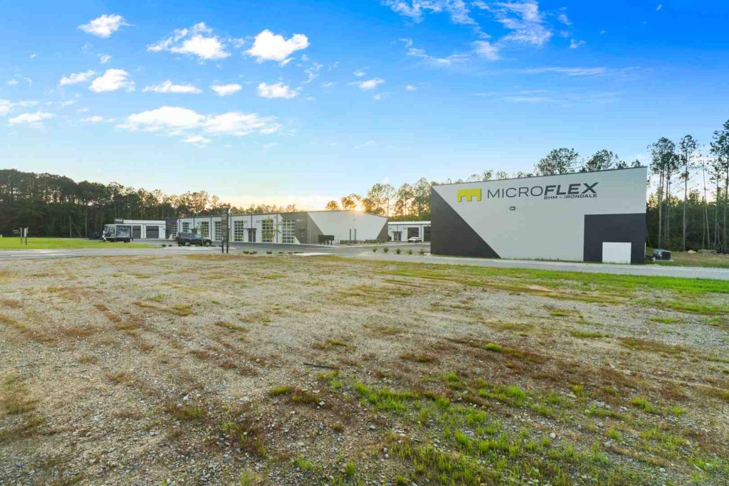 MicroFlex building with angular gray facade and sign, gravel lot, and blue sky above.