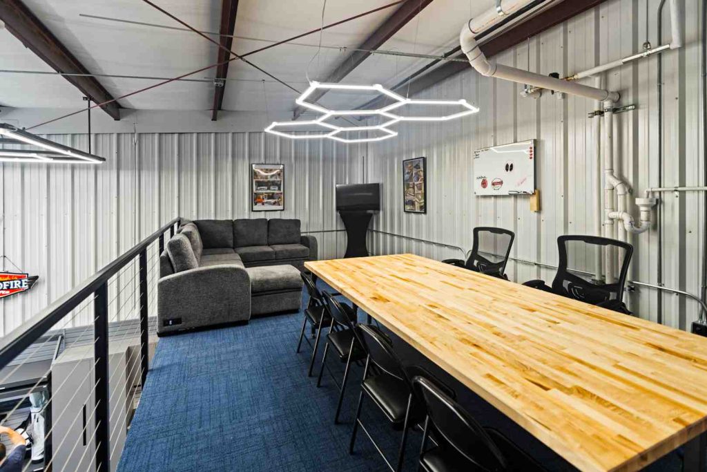 Industrial mezzanine conference area with long wooden table, black chairs, gray sofa, TV, and hexagonal neon lights.