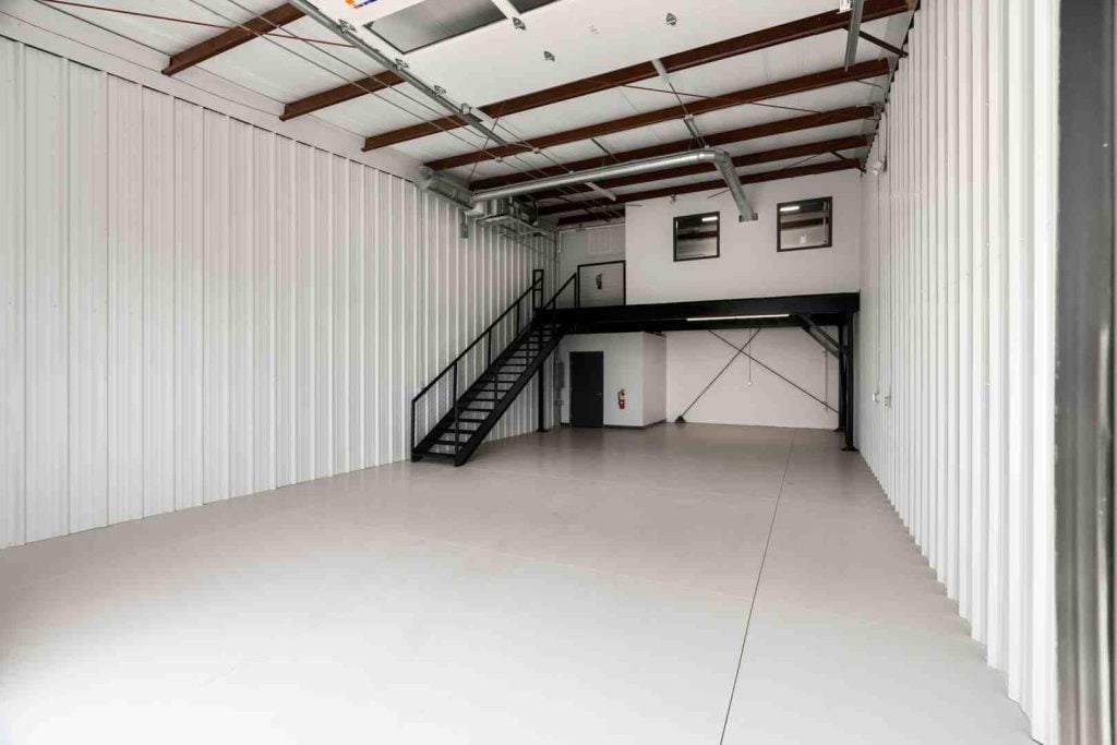 Empty warehouse interior with corrugated metal walls, exposed beams, ducts, and a black staircase to a mezzanine.
