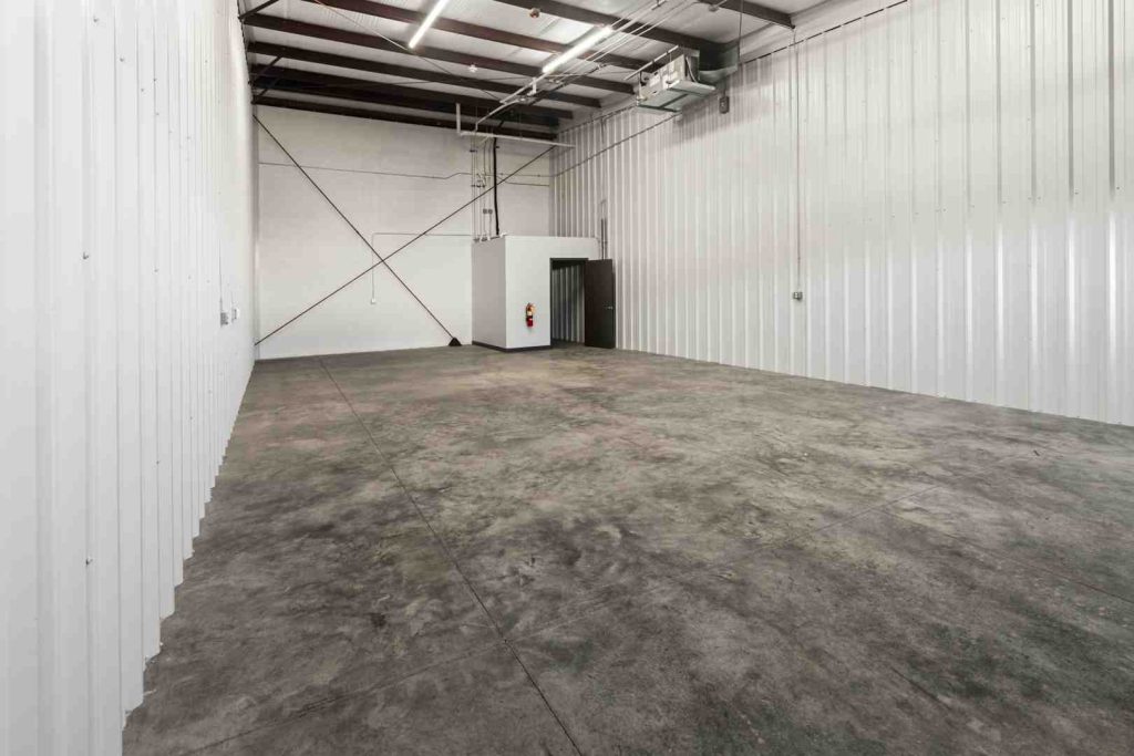 Interior of a white metal-walled warehouse with a concrete floor and a small white utility room with a fire extinguisher.