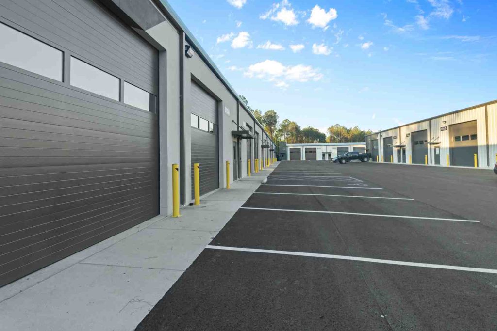 Exterior of a row of gray roll-up warehouse doors, an asphalt parking lot with yellow bollards, under a blue sky.