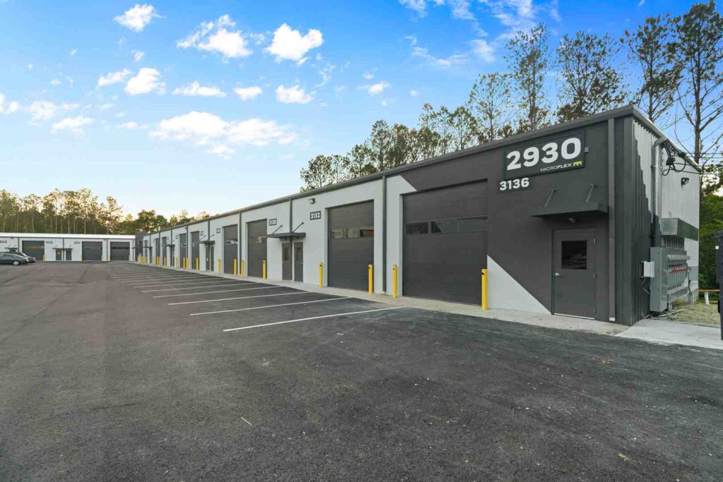 Exterior of a row of warehouse units with gray roll-up doors, yellow safety bollards, and a wide asphalt parking lot.
