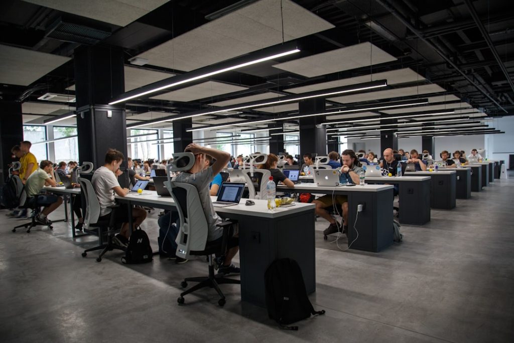 Open-plan office with long rows of desks, employees working on laptops under industrial ceiling lights.