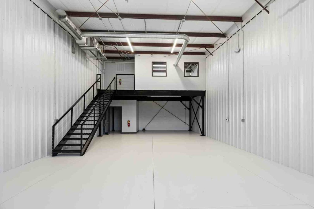 Large empty warehouse interior with a black metal mezzanine and stairs on the left, white corrugated walls, exposed ducts, and bright overhead lighting.