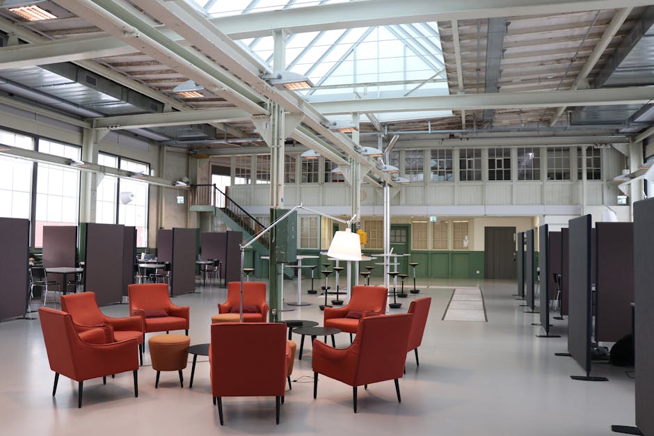 Industrial-style flex space with orange lounge chairs around small tables, gray partitions, mezzanine, and skylit ceiling.