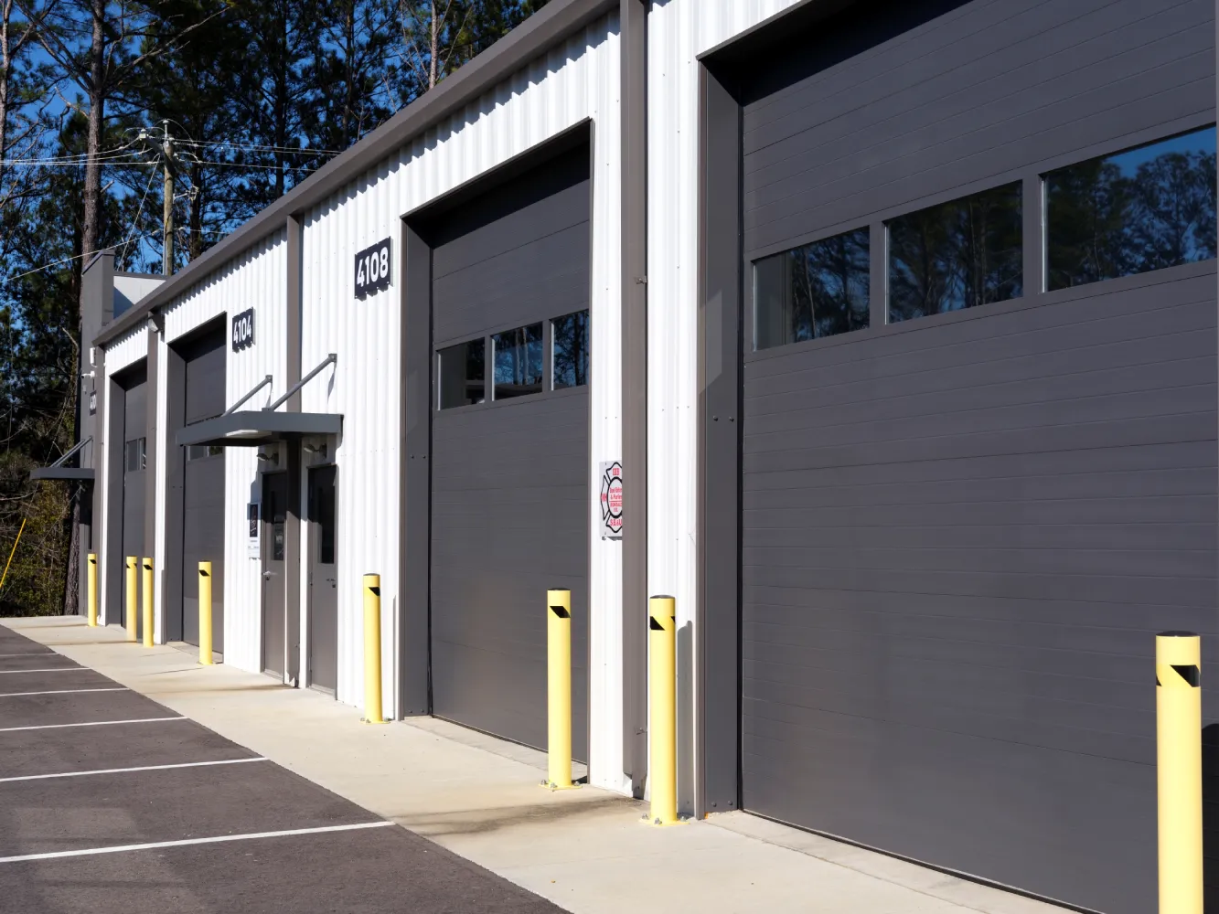Exterior of a row of gray roll-up warehouse doors on white siding, units 4104 and 4108, with yellow bollards and a parking lot.