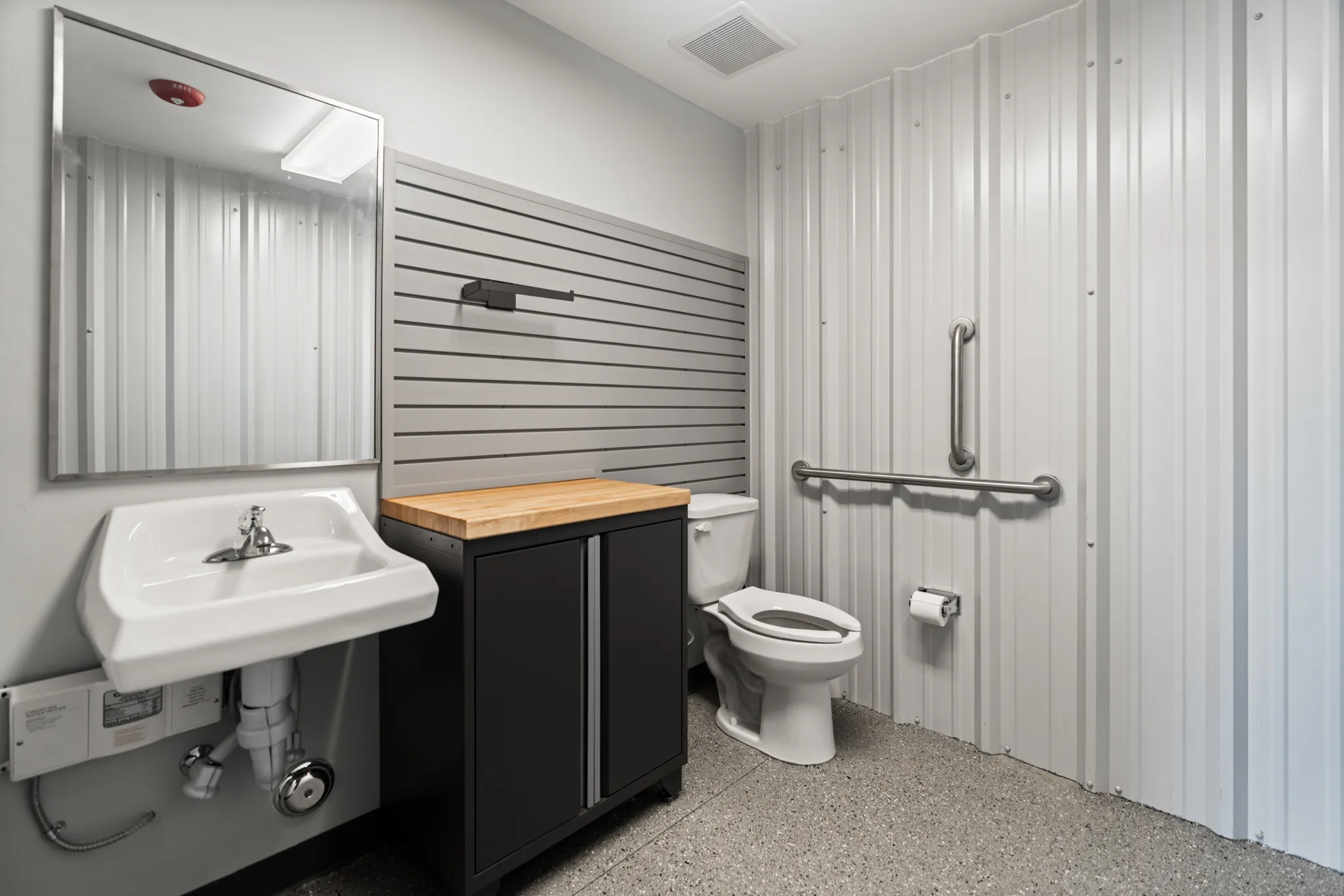 Industrial-style bathroom with wall-mounted sink, cabinet, toilet, mirror, grab bars, and metal walls.