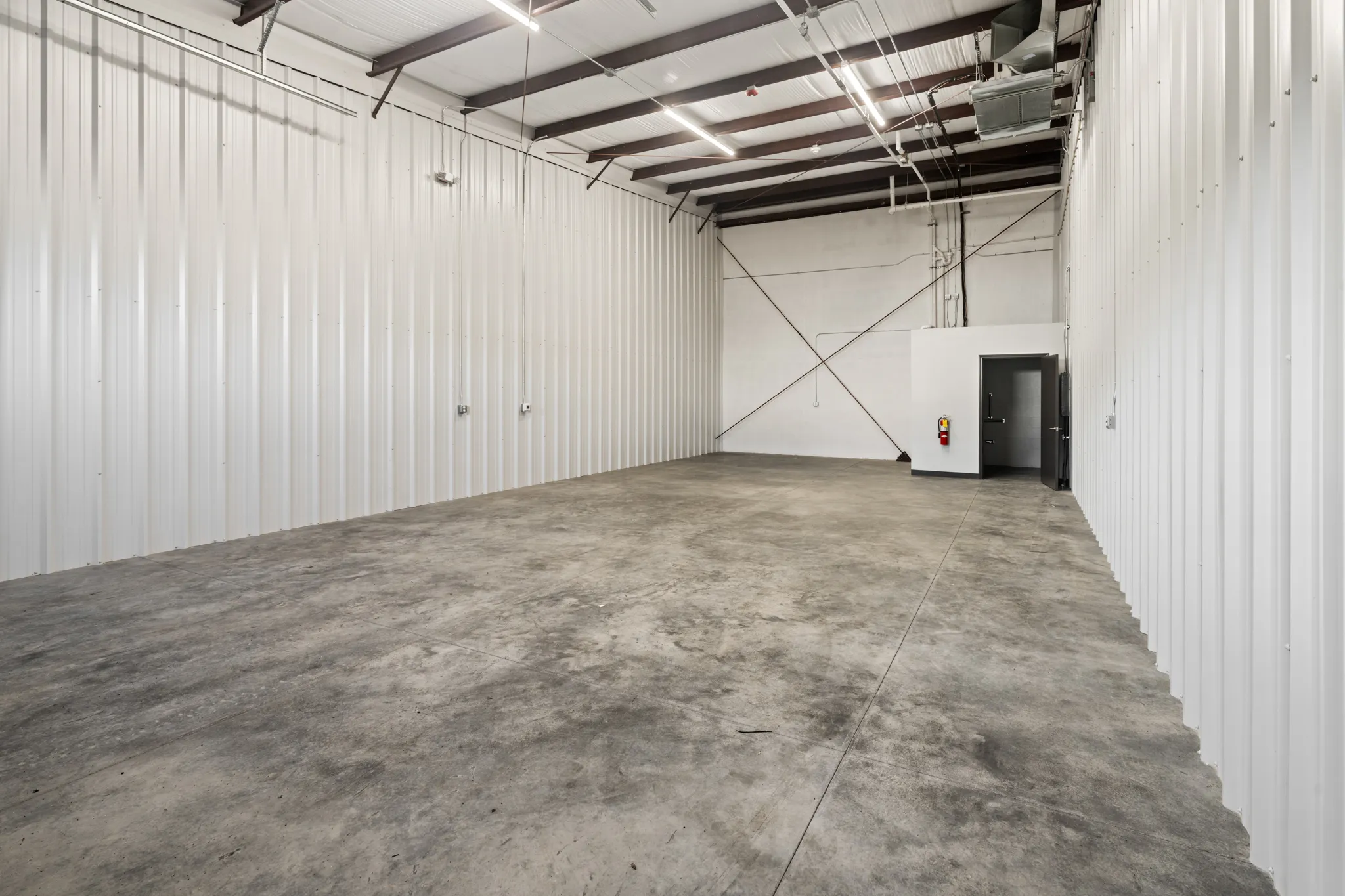 Empty warehouse interior with white corrugated walls, concrete floor, exposed beams, and a small utility room with door.