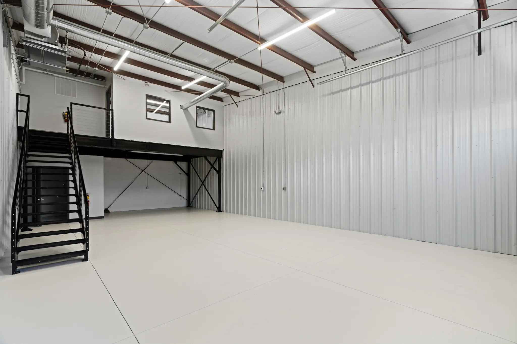 Industrial warehouse interior with black metal staircase to mezzanine, corrugated metal wall, and exposed ducts.