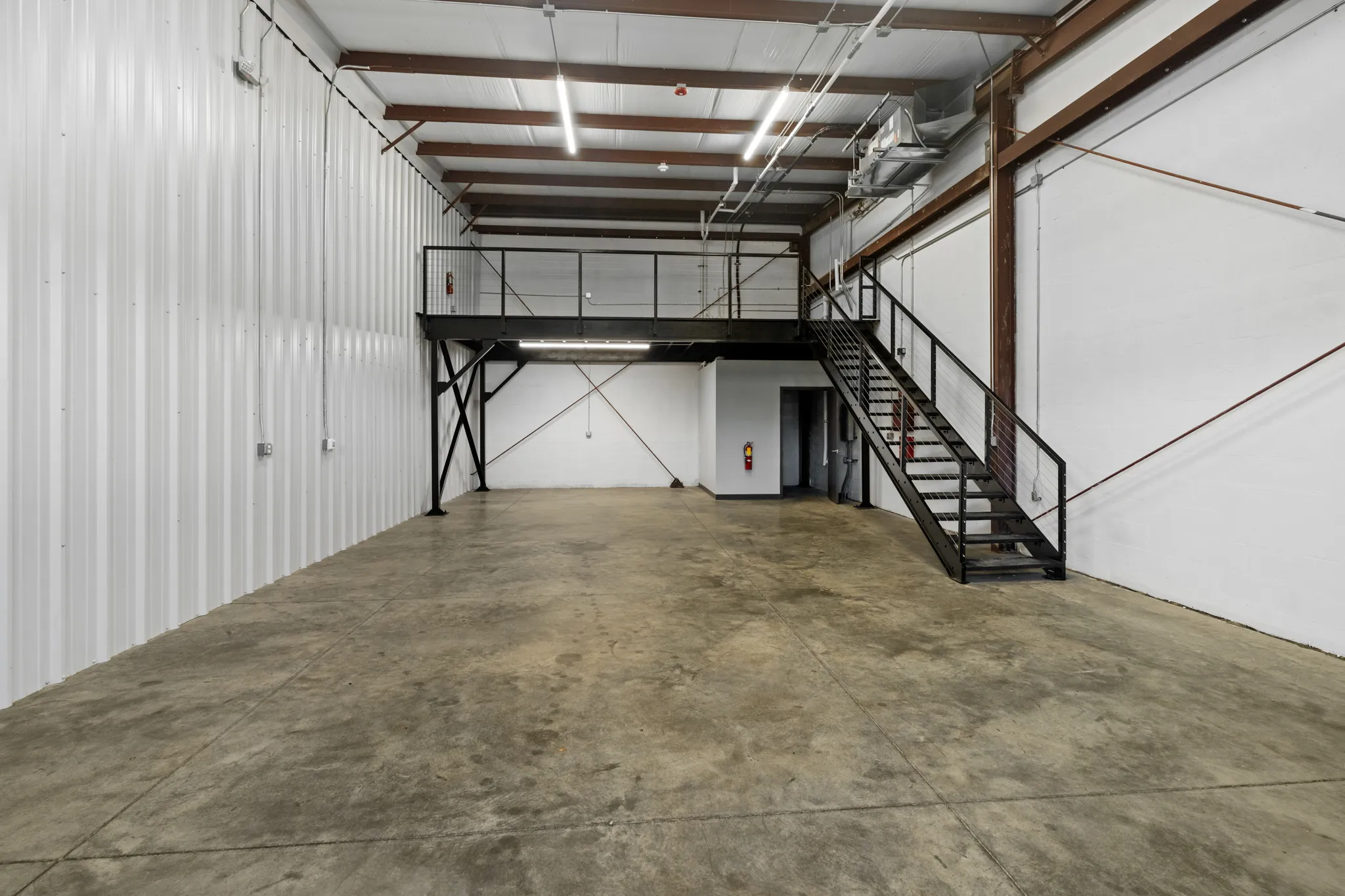 Industrial warehouse interior with concrete floor, white corrugated walls, and a black steel mezzanine with staircase.