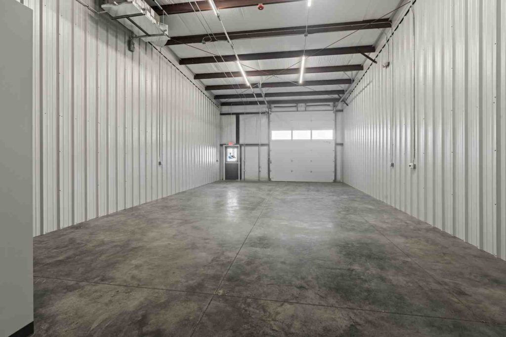 Empty industrial warehouse interior with white corrugated metal walls, concrete floor, exposed beams, and a roll-up door at the far end.
