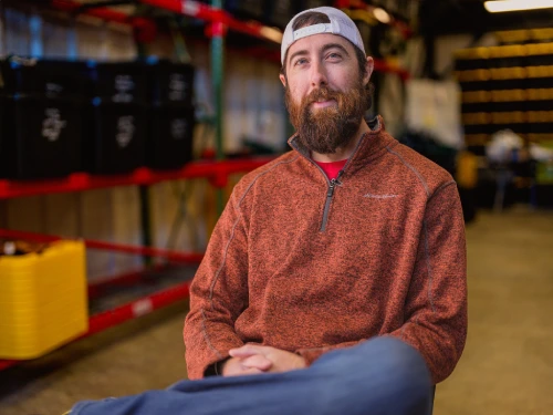 Bearded man in a gray cap and orange pullover sits in a warehouse with red metal shelving and a yellow crate.