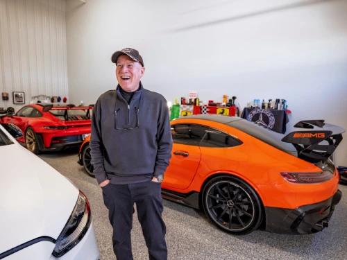 Smiling man in a dark hoodie and cap stands in a car showroom beside an orange sports car and other vehicles.