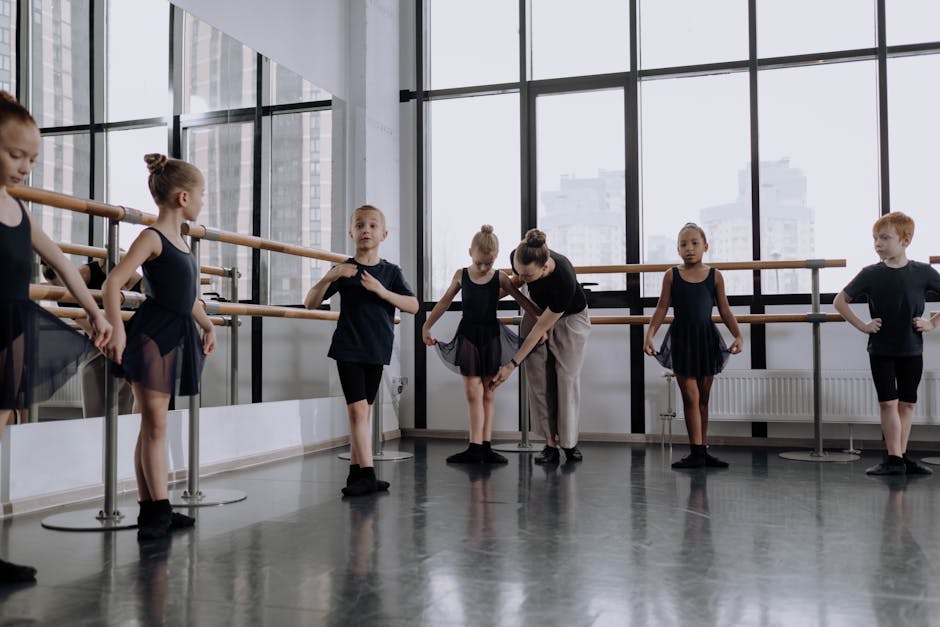 Young ballet students in black leotards practice at a barre in a bright studio with large windows; instructor assists.